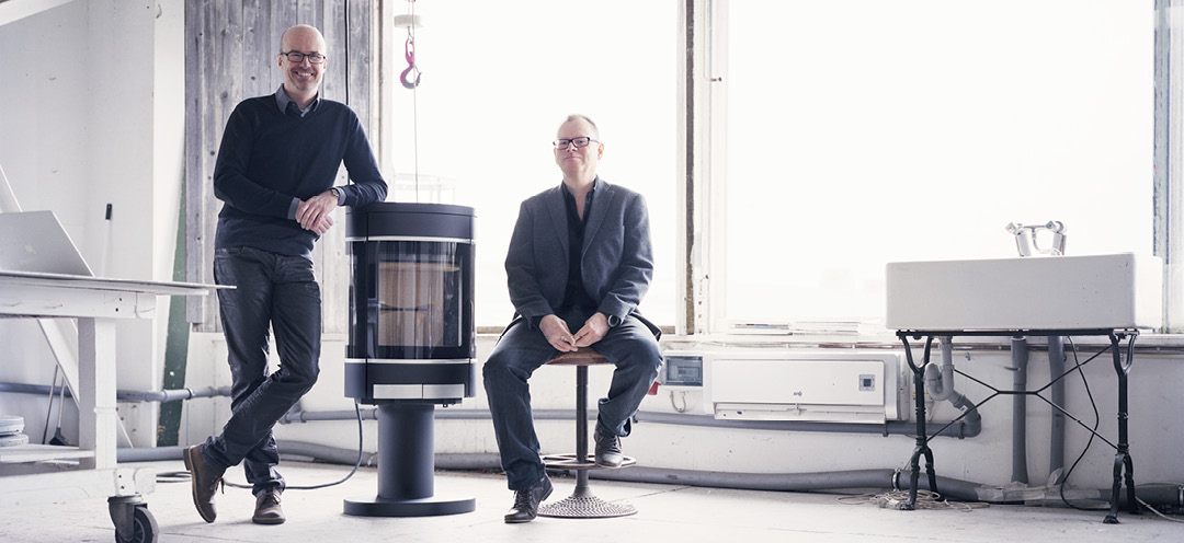 Two middle-aged men in a bright factory room, one leaning against a black fireplace Scan 68 and the other sitting on a stool