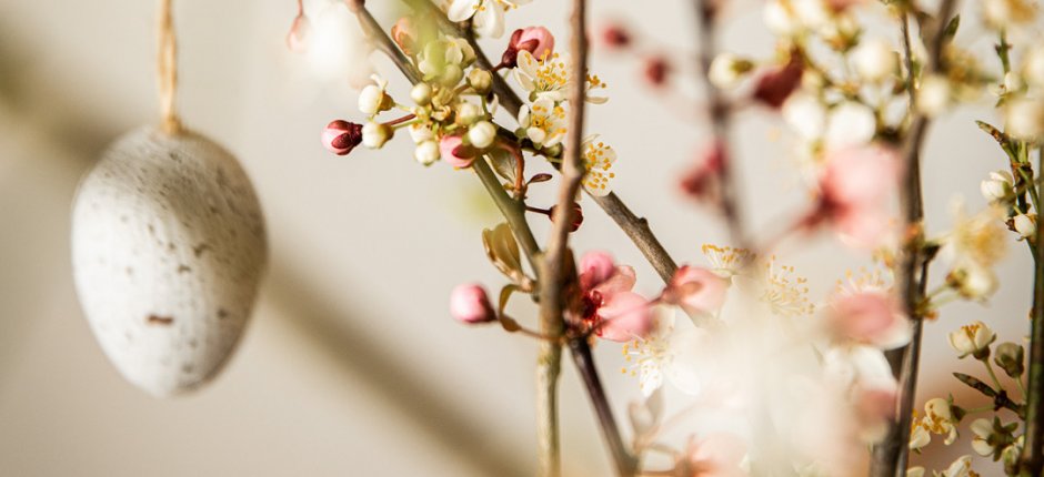 Small eggs painted white with brown dots hanging on a branch with flowers