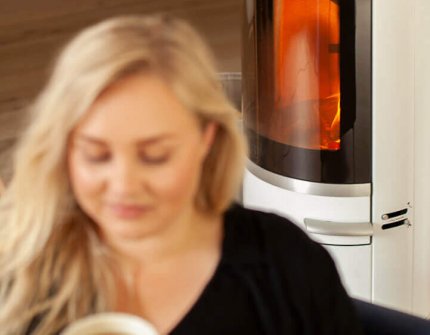 Blonde woman sitting in front of a white wood-burning stove Scan 83-2 with a cup of coffee in her hands