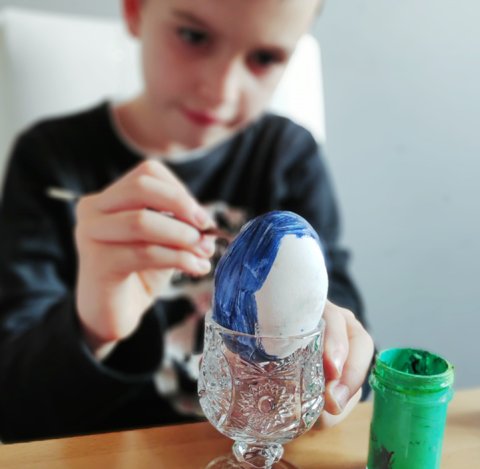 Boy sitting at a table and painting Easter eggs with a blue color paint