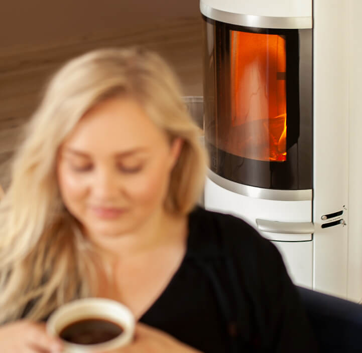 Blond woman sits in front of a white Scan 83-2 wood stove with a cup of tea in her hands