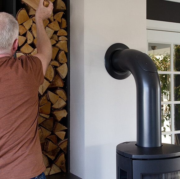 Black wood stove with knee pipe into white pipe. Man in brown t-shirt puts firewood in wood niche.