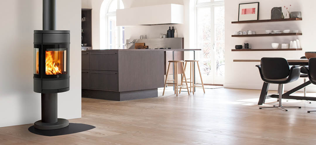 Oval, black wood-burning stove with side glass Scan 68-9 on column placed in a bright kitchen with modern brown interior