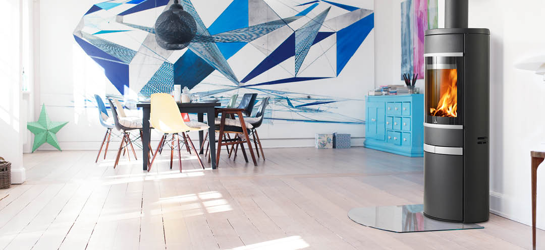 Black oval wood-burning stove with side glass, aluminum handle and decorative strips Scan 68-12 in a white living room with large graphic art above the dining room