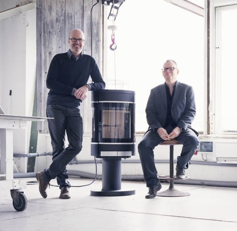 Two elderly male product developers are leaning against a black wood stove on a column Scan 68-10